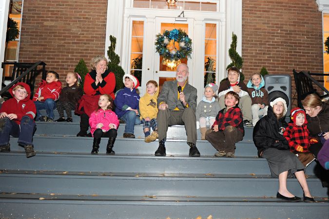 A group of young children sit on the steps of a porch, surrounding a man who is holding a microphone and singing carols. Behind them is a glass door with white trim holding a wreath.