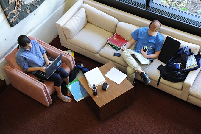 Aerial shot of two students studying separately, one in a reddish chair and one on a long, off-white couch.
