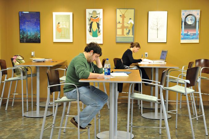 Three high tables and chairs, a wall of art in the background, and two students at separate tables. The walls are a mustard yellow and feature various vertical artworks.