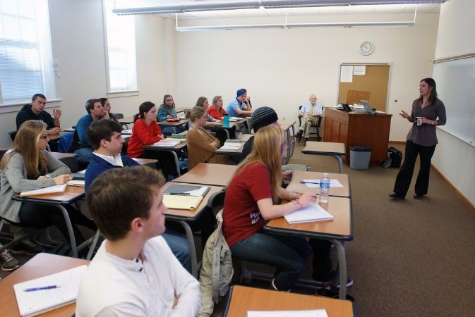 Students sit at two-person desks in front of a whiteboard and speaker, with a desk in the left of the classroom.