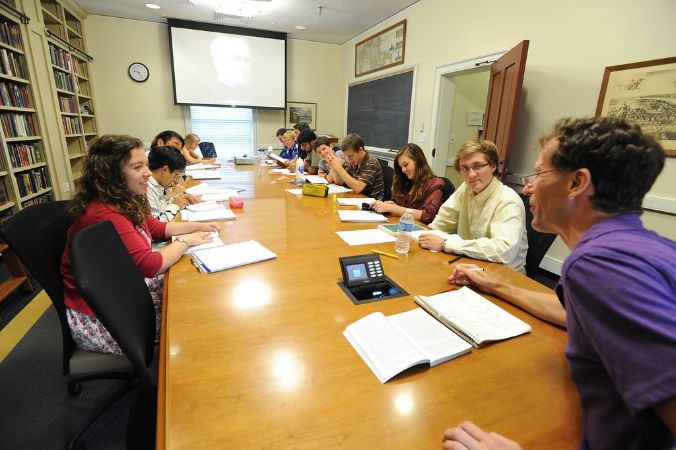 Students sitting at a long table looking at a projection, bookshelves on one side of them.