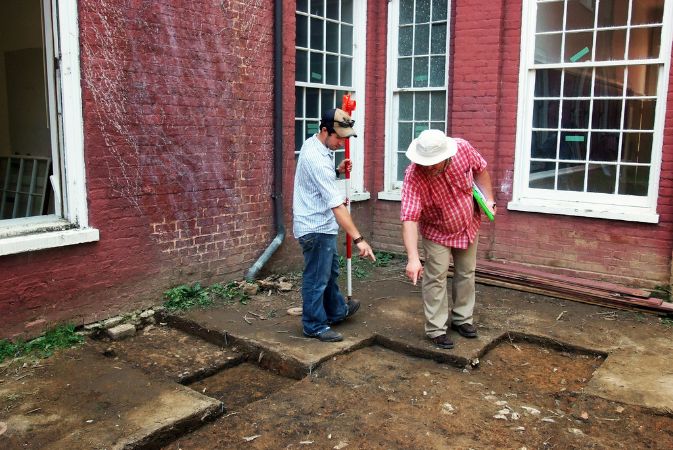 In a square area between two red brick walls and windows, two workers observe the recently excavated dirt area between the buildings.