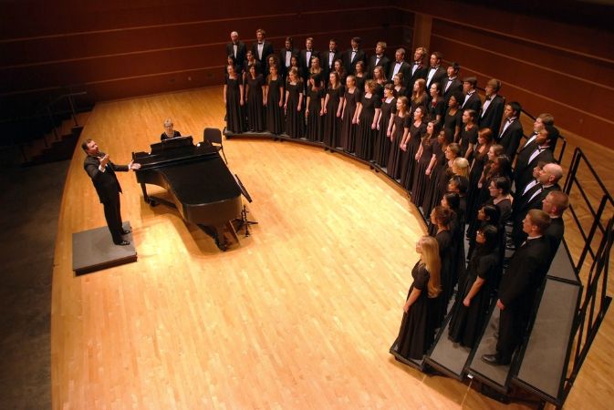 The choral director conducts from a light wooden stage next to a piano, across from men and women singers on rounded risers.